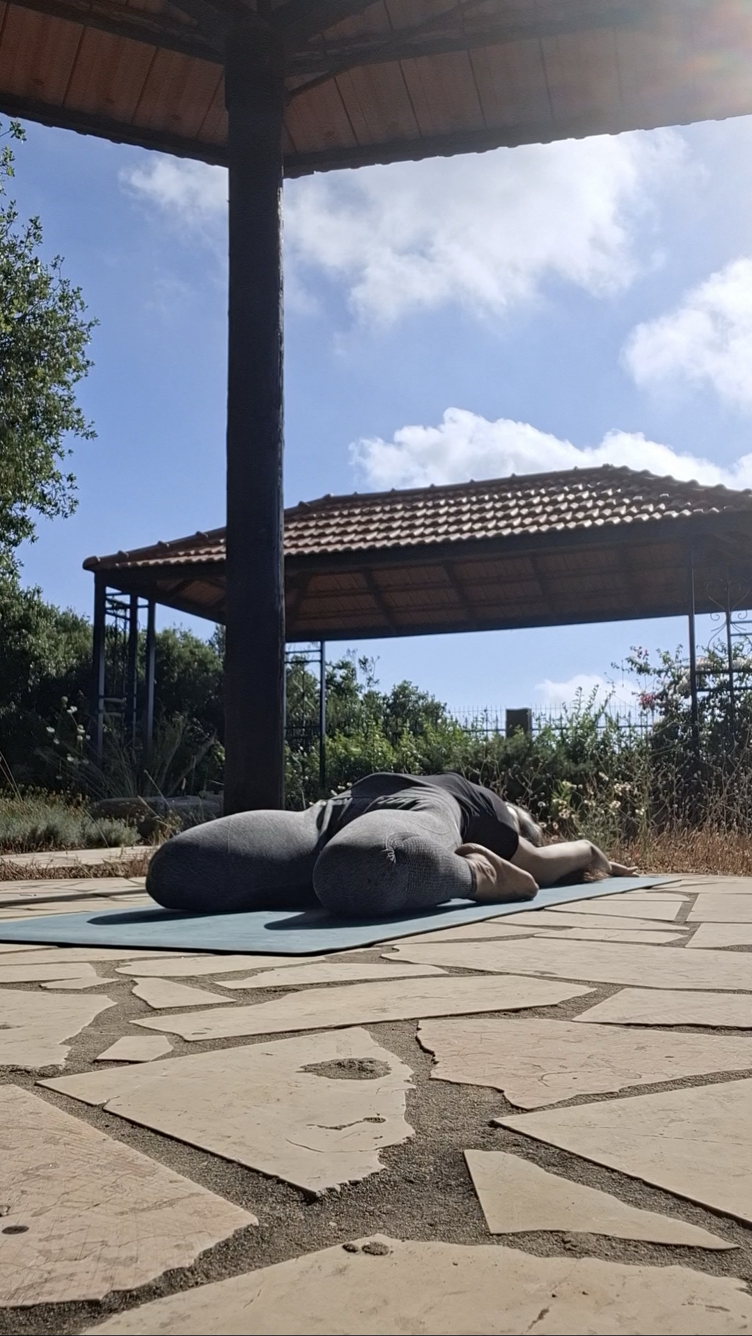 A student resting in a supported forward fold on a mat under a wooden pergola in the hills